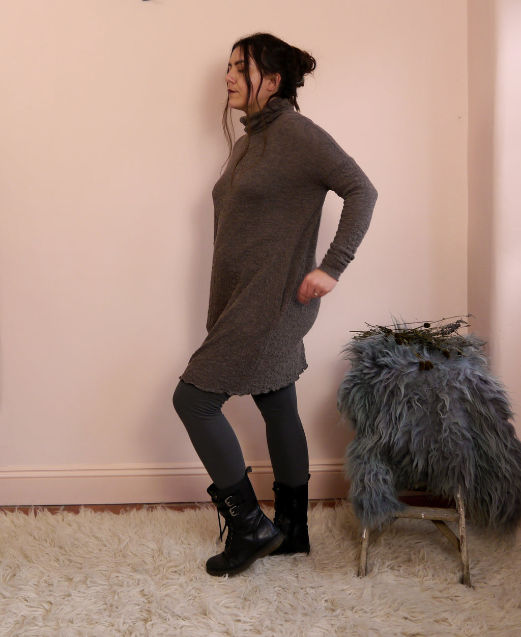 Woman wearing a long-sleeve brown jumper dress with a turtleneck, standing next to a small stool with a fur cover against a pink wall.