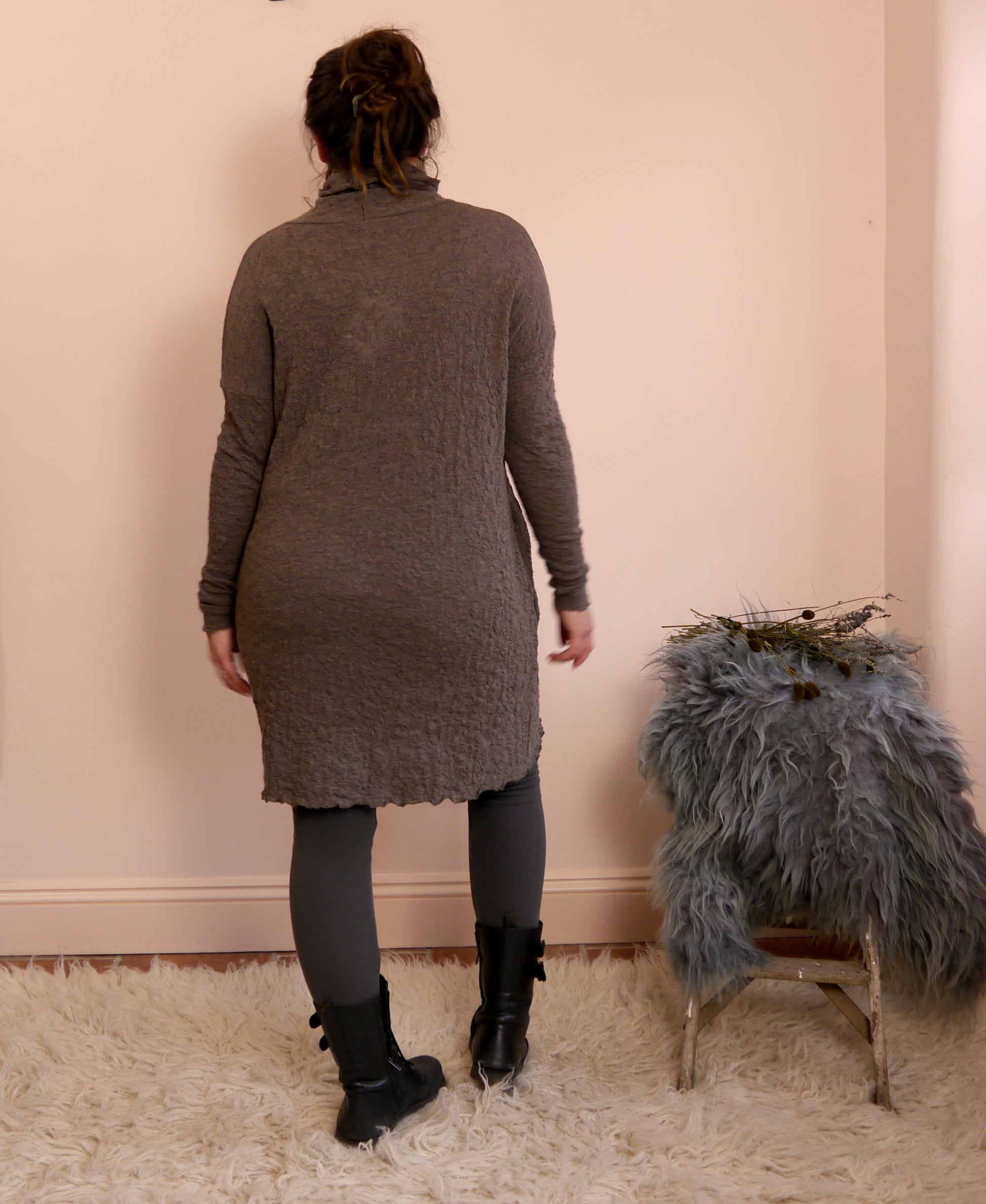 Person wearing a brown merino wool turtleneck dress standing in front of a pink wall with a small stool and decorative items.