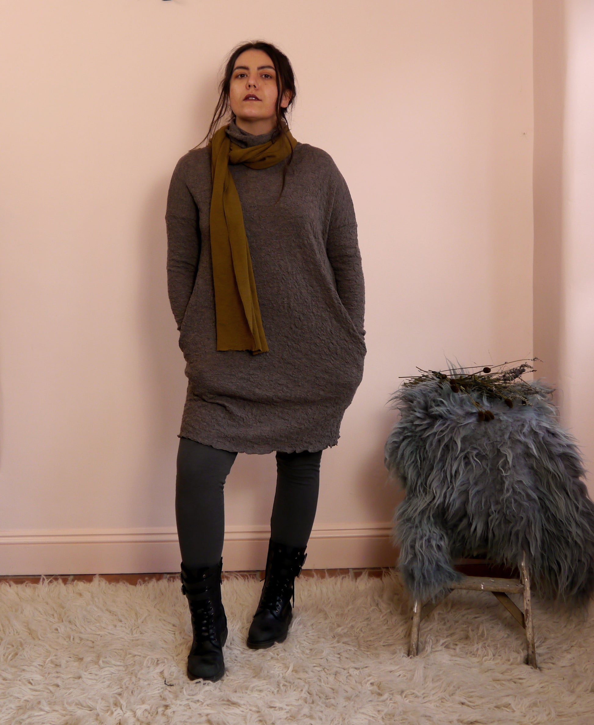 Woman wearing a brown merino wool turtleneck dress and bronze scarf standing against a pink wall with a fur-covered stool.