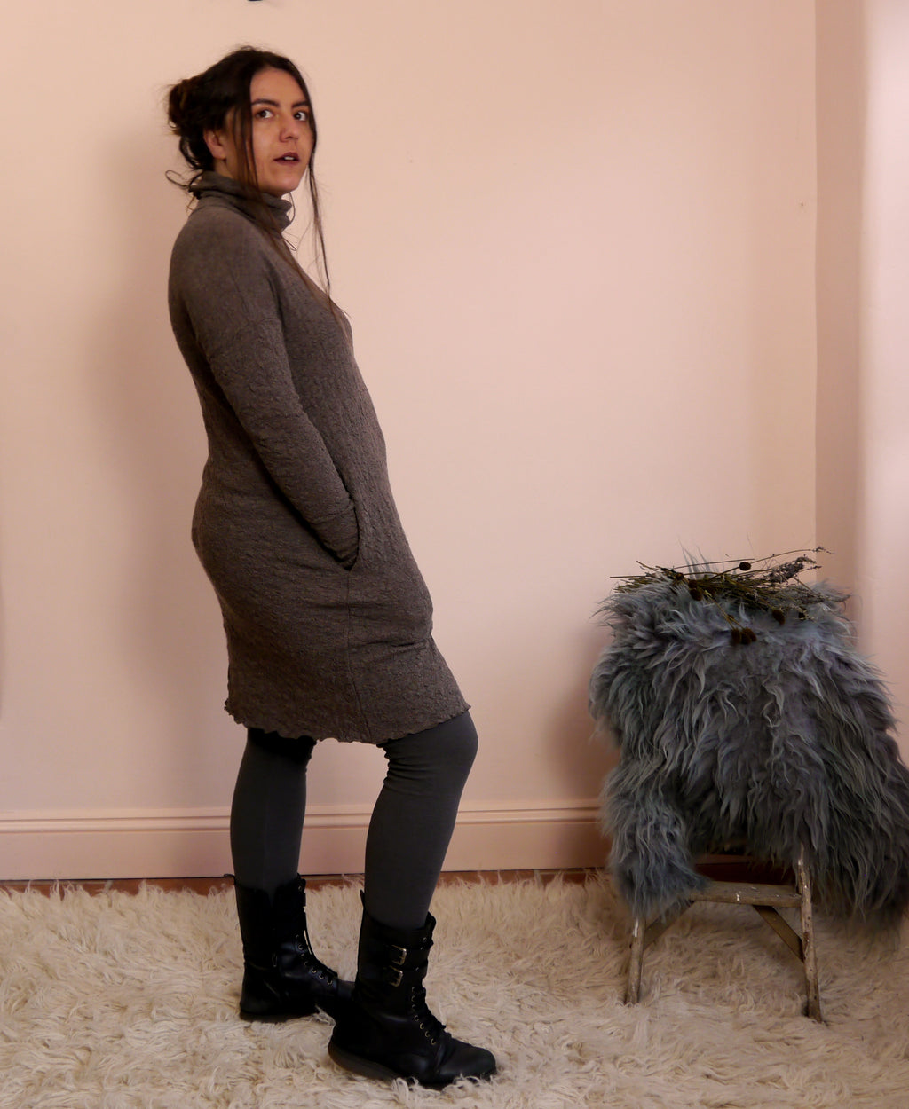 Woman wearing a brown merino wool turtleneck dress standing next to a chair with a fur cover against a pink wall.