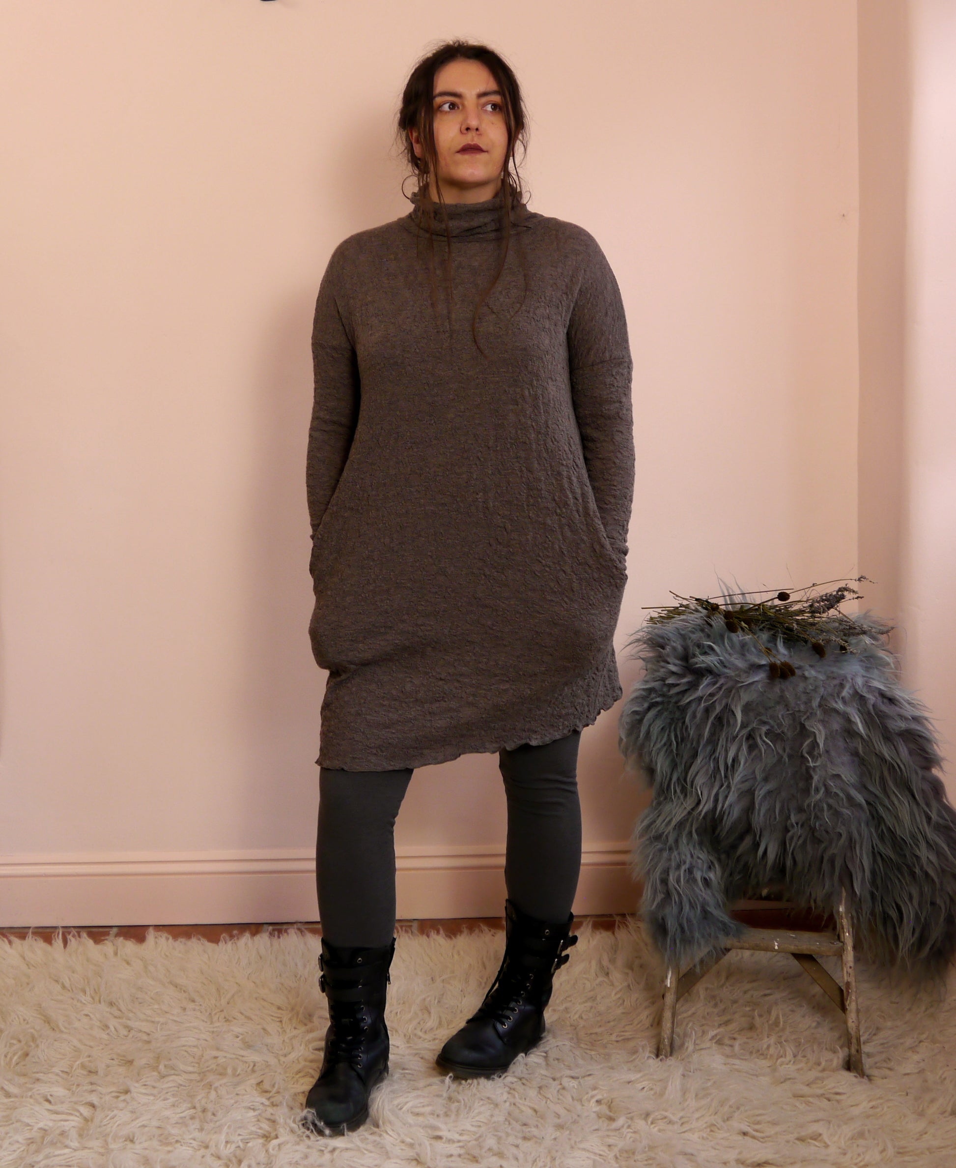 Woman wearing a brown merino wool turtleneck dress standing next to a small table with a fur cover against a beige wall.