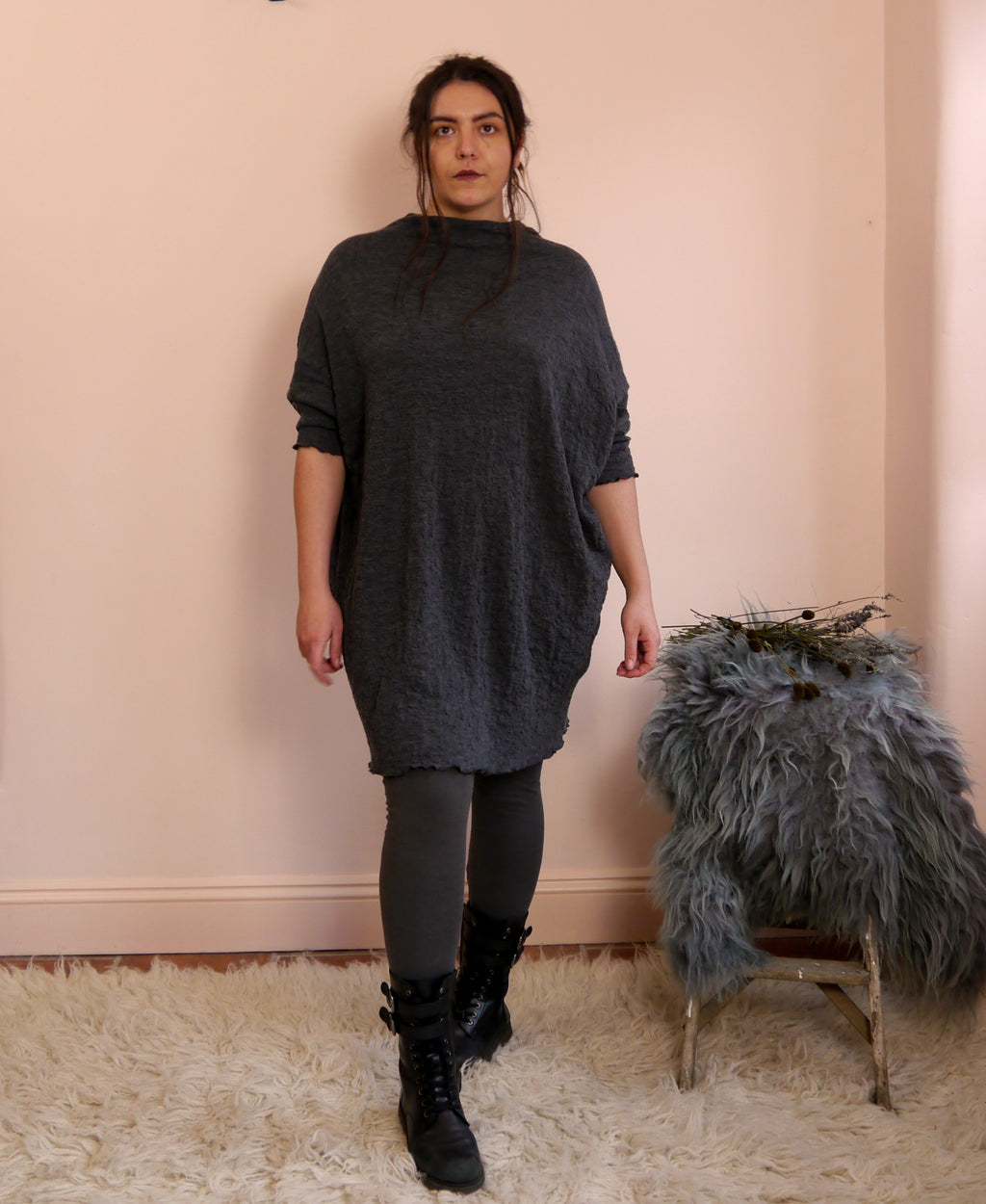 Person wearing a dark gray merino wool dress standing next to a small stool with a fur cover against a pink wall.