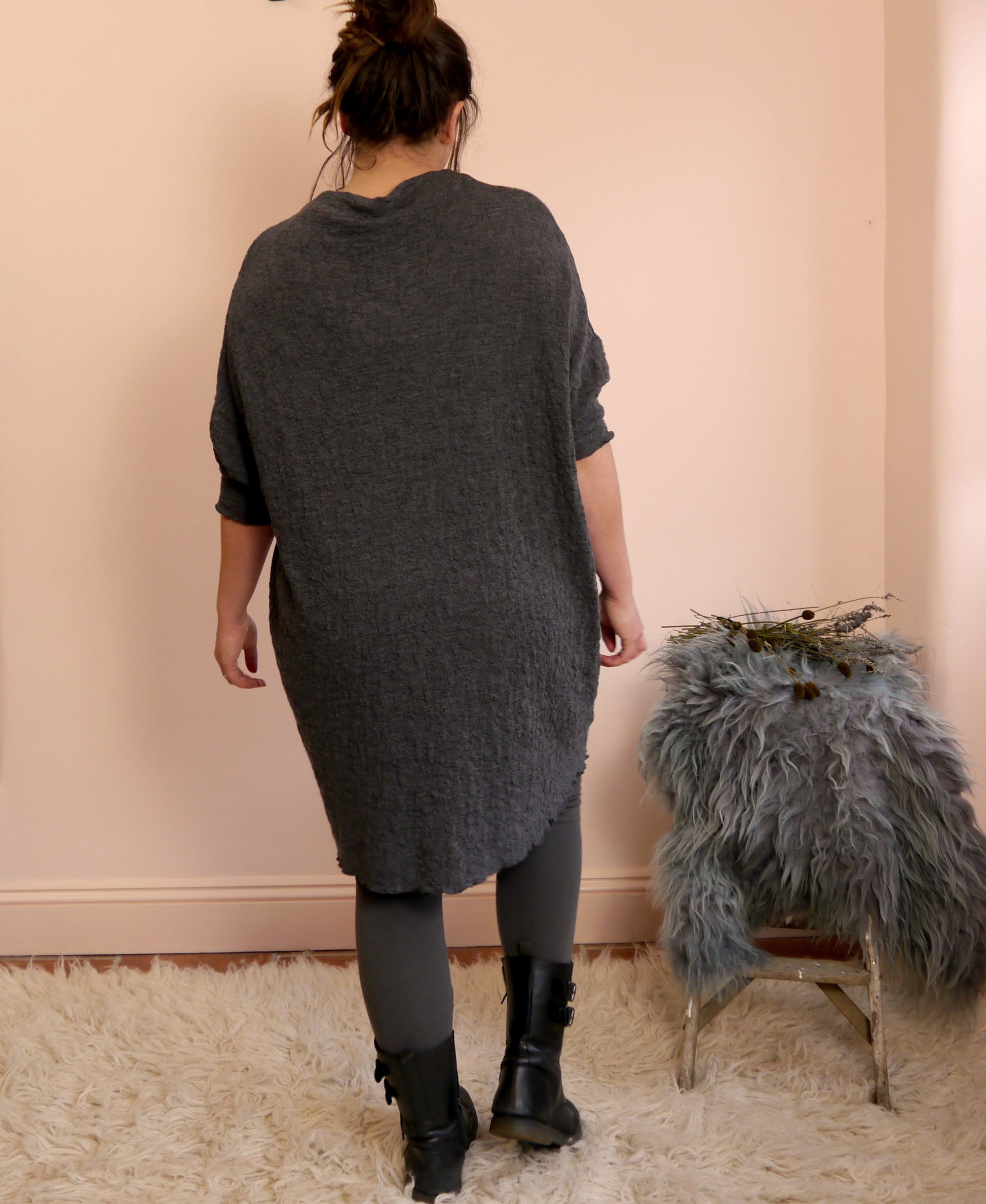 Person wearing a dark gray merino wool dress standing in a room with a pink wall and a fur-covered chair.