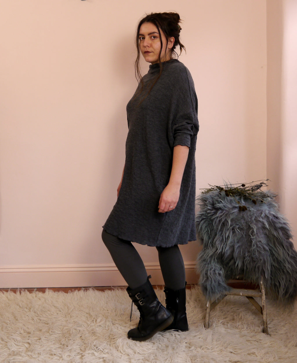 Woman wearing a dark gray merino wool dress standing against a light pink wall with a textured rug and chair.