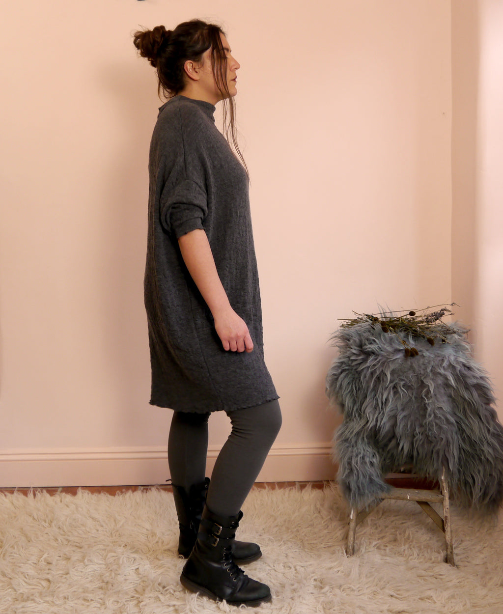 Woman wearing a ldark gray merino wool dress standing against a light pink wall with a small stool and decorative items.