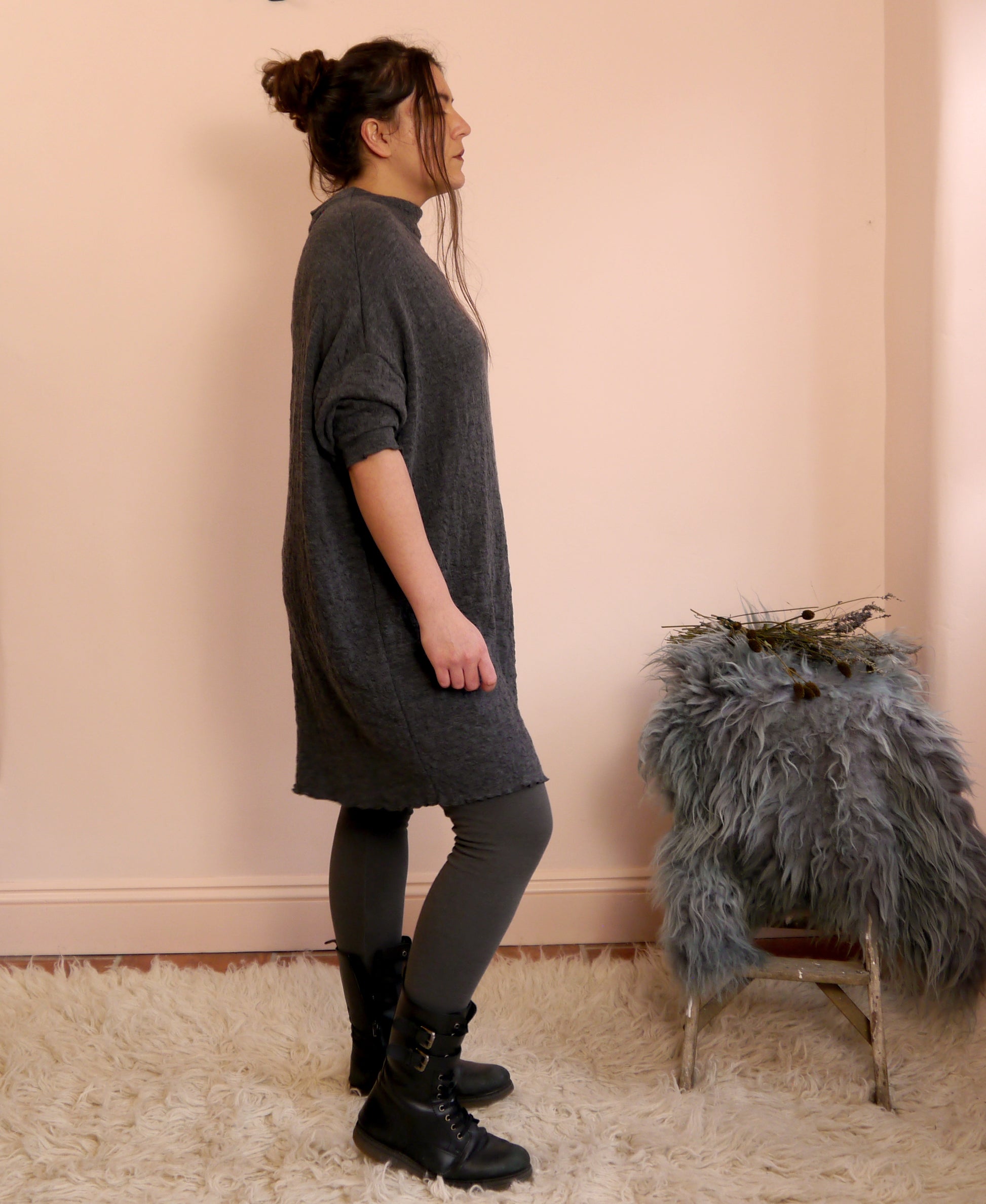 Woman wearing a ldark gray merino wool dress standing against a light pink wall with a small stool and decorative items.