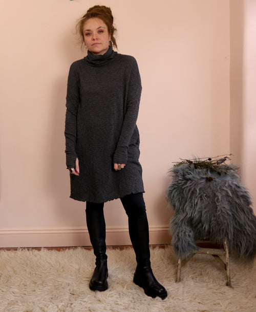 Woman wearing a dark gray merino wool turtleneck dress standing against a pink wall with a small stool and decorative item.