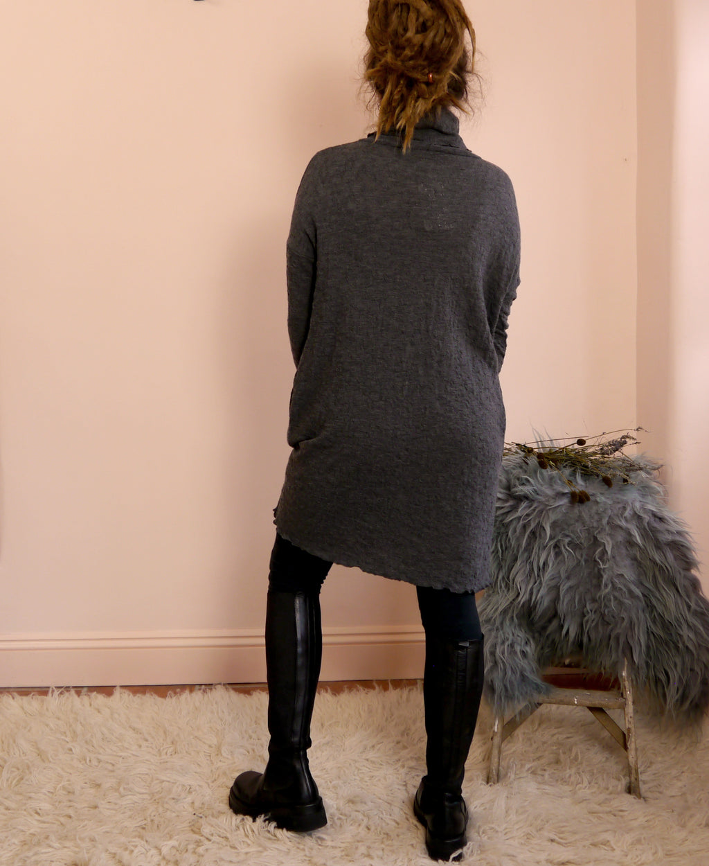 Person wearing a dark gray merino wool turtleneck dress standing in front of a pink wall with a chair and decorative items.
