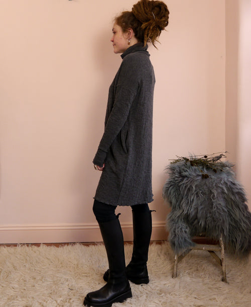 Woman wearing a dark gray merino wool turtleneck dress standing against a pink wall with a fluffy rug and chair.