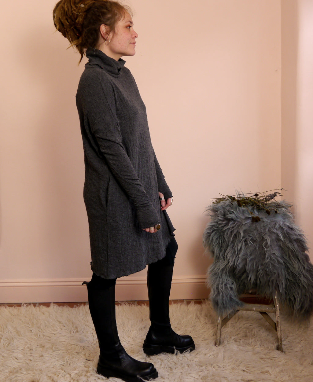 Woman wearing a long dark gray merino wool turtleneck dress standing against a light pink wall with a small stool and decorative items.