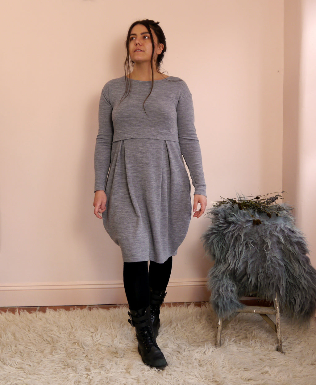 Woman wearing a gray merino wool dress standing next to a small stool with a fur cover against a pink wall.
