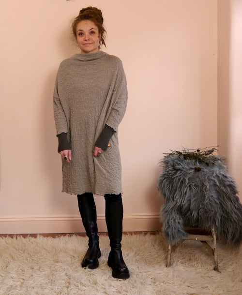 Woman wearing a taupe merino wool dress standing against a pink wall with a textured rug and fur-covered stool.