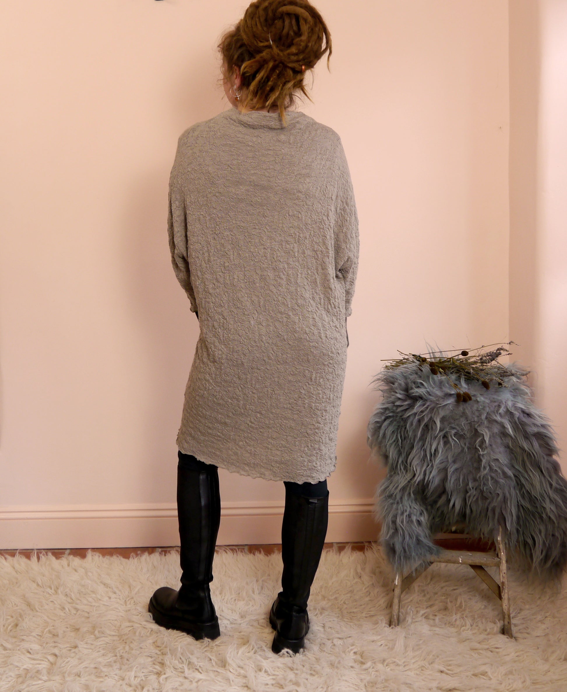 Person wearing a merino wool dress standing in a room with a pink wall and a fur-covered chair.