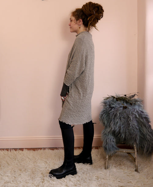 Woman in a merino wool dress standing next to a small stool with fur against a pink wall.