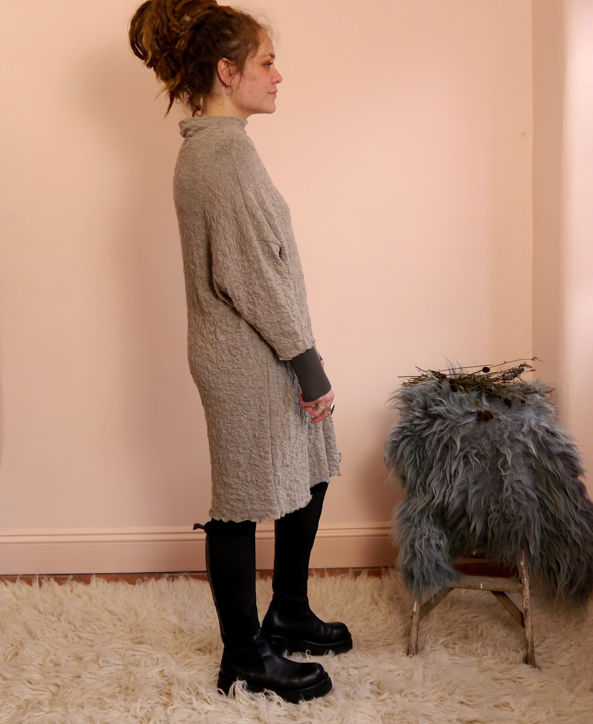 Woman wearing a taupe merino wool dress standing next to a chair with a fur cover against a pink wall.